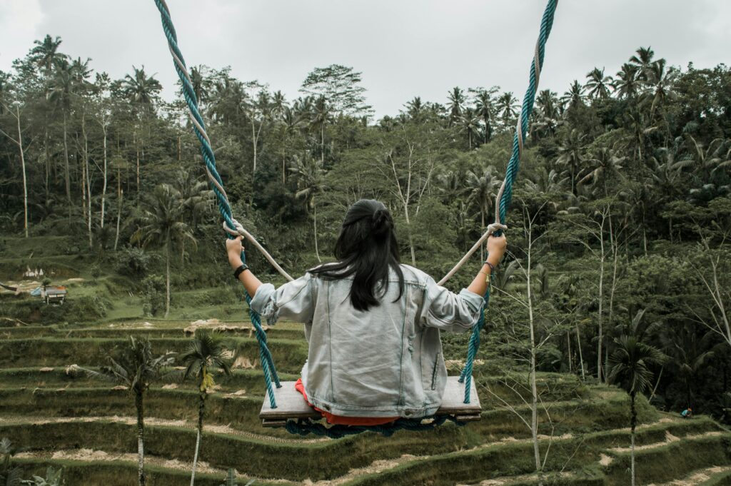 A woman in a denim jacket enjoys a swing with a scenic view of the lush rice terraces in Ubud, Bali.